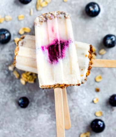 Top view of three vegan blueberry popsicles stacked on a grey blue background with blueberries on the side