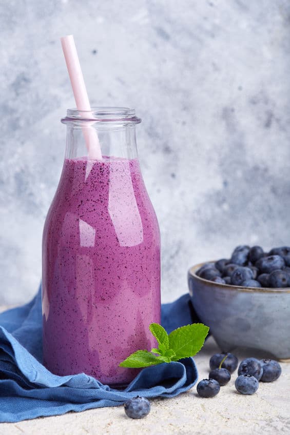 Side view of a healthy blueberry smoothie in a glass jar with a straw topped with a bowl of fresh blueberries on the side
