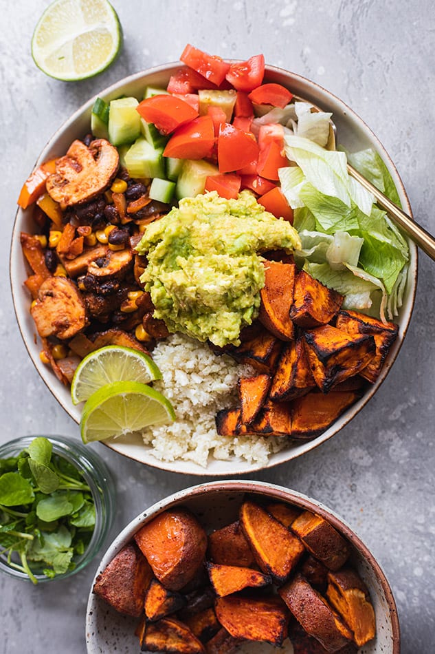 An assembled vegan veggie bowl beside half a lime and a bowl of roasted sweet potatoes