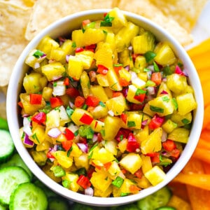 Top close-up shot of a serving of fresh pineapple salsa in a white bowl with sliced cucumbers, bell peppers and tortilla chips