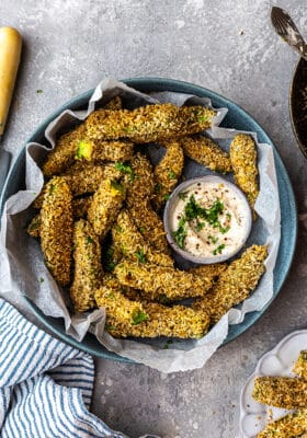 Top view of baked avocado fries in a blue bowl with avocado dip on a grey background