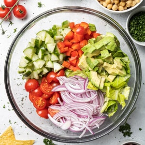 Overhead view of chopped cucumber, tomatoes, bell peppers, avocado and red onions side-by-side in a large clear mixing bowl to make a Mediterranean Chickpea Salad
