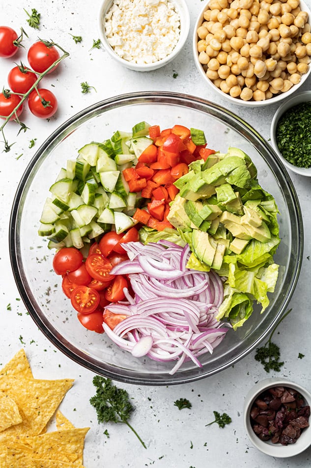 Chopped cucumber, cherry tomatoes, bell peppers, avocado and red onions side-by-side in a large glass mixing bowl
