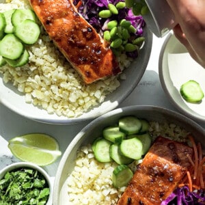Two maple miso salmon fillets in two white bowls with cauliflower rice, edamame, cucumbers, carrots and red cabbage