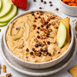 Overhead view of pumpkin dip in a bowl with mini chocolate chips, chopped nuts, and an apple slice