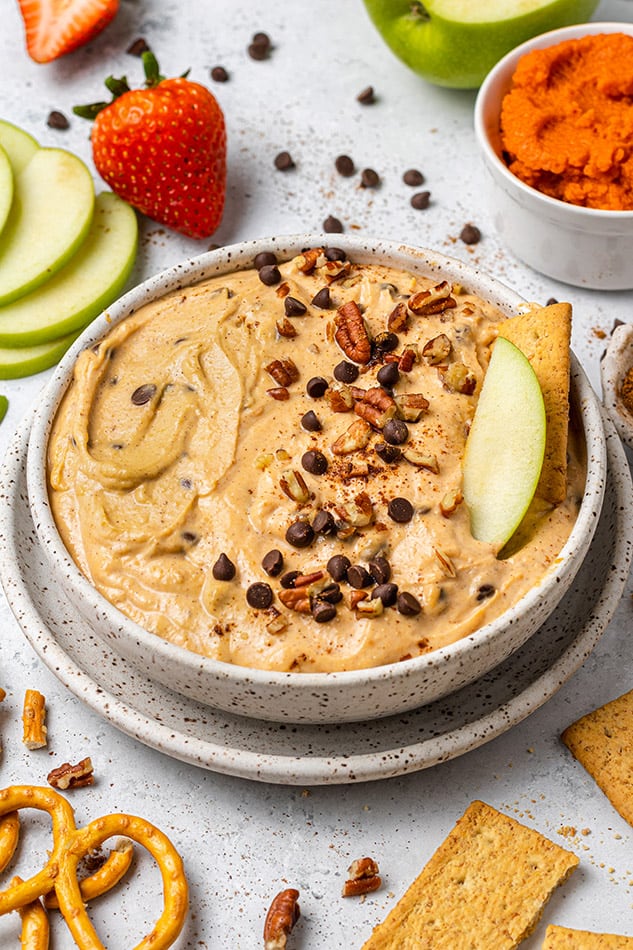 A bowl of pumpkin dip with mini chocolate chips and chopped nuts with an apple slice and graham cracker 