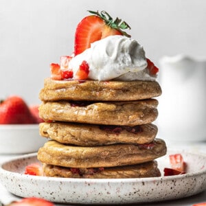 Side shot of a stack of five fluffy strawberry pancakes topped with sliced and diced strawberries with whipped cream on a white plate