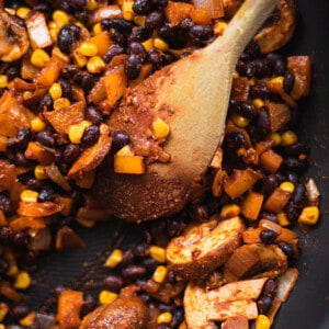 Black beans, mushrooms, peppers and corn cooking in a skillet while being stirred with a wooden spoon