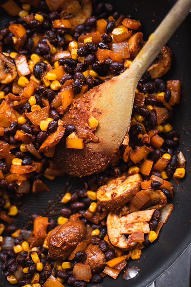 Black beans, mushrooms, peppers and corn cooking in a skillet while being stirred with a wooden spoon