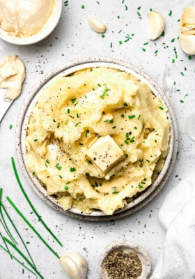 A bowl of creamy vegan mashed potatoes on a countertop beside garlic cloves and fresh chives