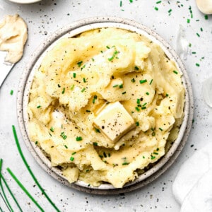 A bowl of creamy vegan mashed potatoes on a countertop beside garlic cloves and fresh chives