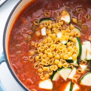 Vegetable broth, diced tomatoes, zucchini and dried pasta shells in a blue pot