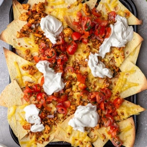 Overhead shot of loaded vegan nachos layered with seasoned tofu crumble, vegan sour cream and fresh tomato salsa in a baking pan on a grey background