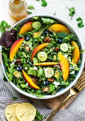 Top view of arugula salad with peaches and blueberries in a white bowl on a wooden cutting board on a grey background