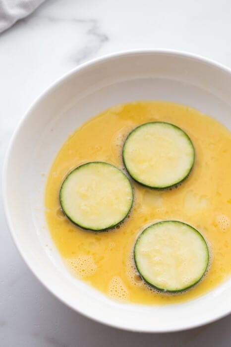 Three sliced zucchini rounds in a bowl of beaten eggs