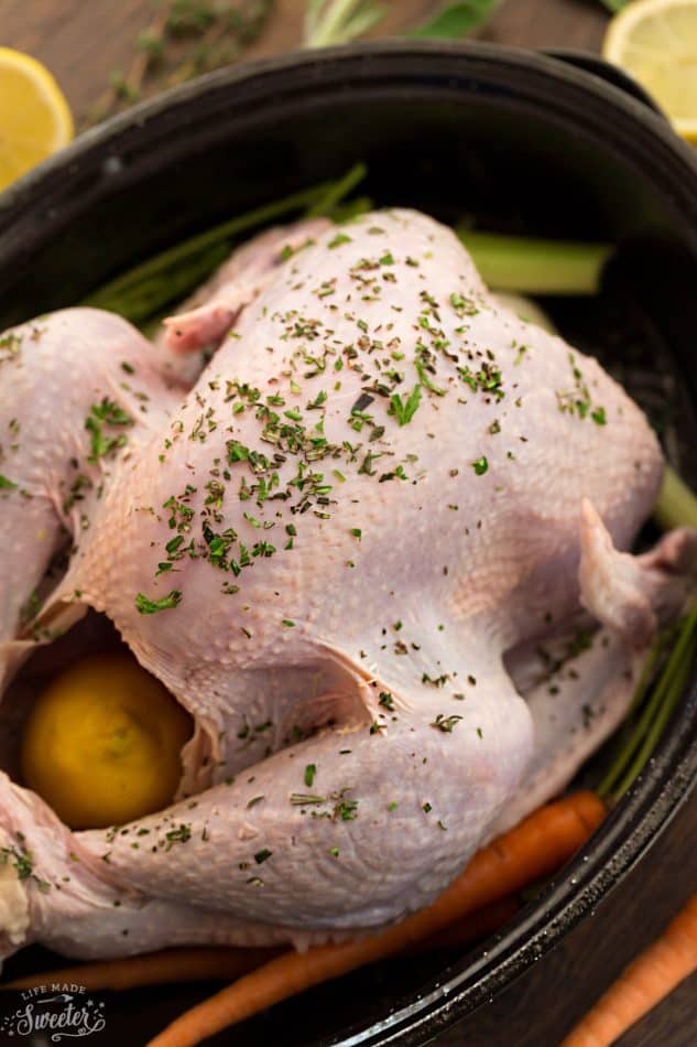 Top view of a raw whole turkey in a black roasting pan with carrots, lemon and herbs