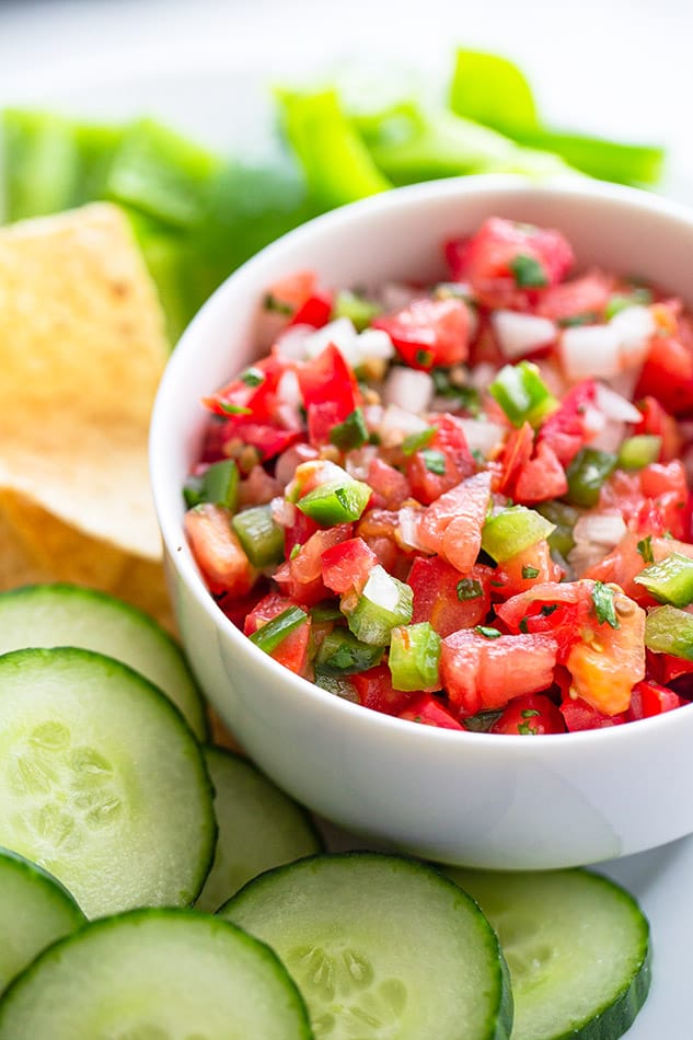 Side shot of a bowl of homemade tomato salsa in a white bowl with cucumber slices and tortilla chips