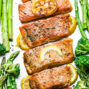 Close up of roasted lemon butter salmon fillets on a baking sheet with lemon slices and roasted veggies on the side