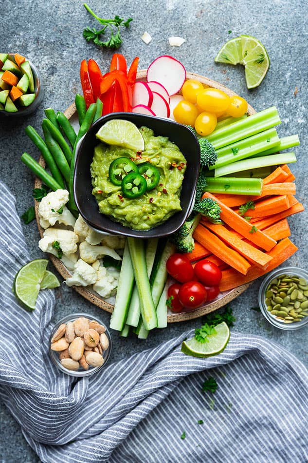 A round serving dish full of veggies, with a bowl of guacamole, next to a bowl of pistachios 