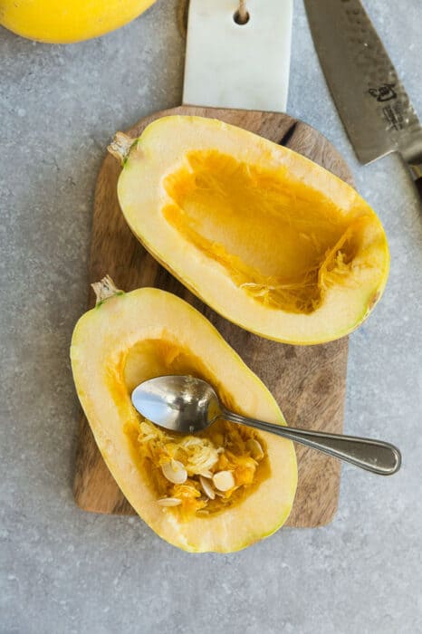 A spoon scooping seeds out of two raw spaghetti squash halves on a wooden cutting board