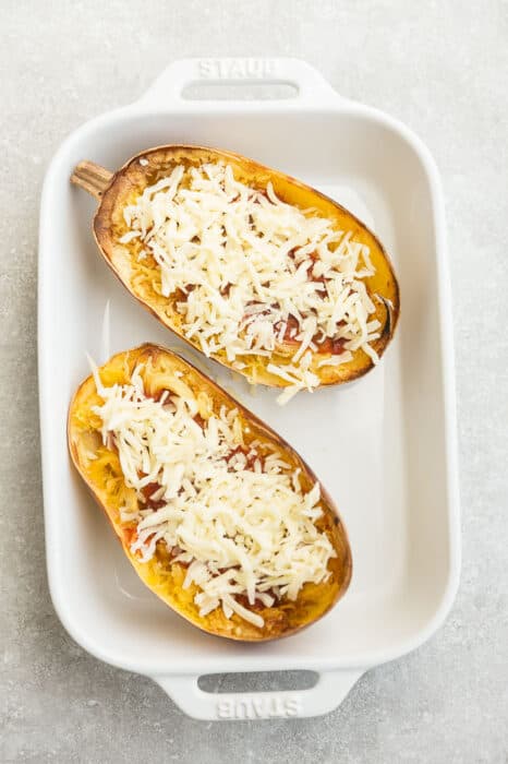 Overhead view of spaghetti squash halves with tomato sauce and cheese in a baking dish