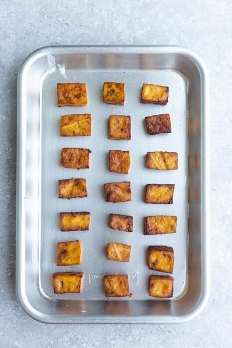 Crispy baked tofu cubes lined up on a metal baking sheet with about an inch of space between each one