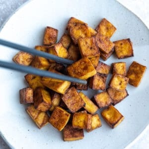 Crispy cooked tofu cubes piled onto a white plate with a pair of chopsticks grabbing one of the cubes