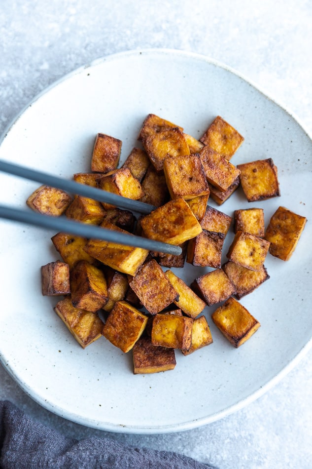 Crispy cooked tofu cubes piled onto a white plate with a pair of chopsticks grabbing one of the cubes