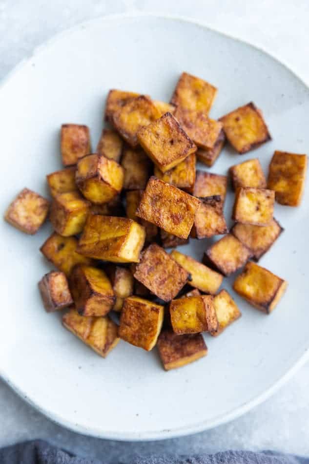A pile of crispy baked tofu on a large white plate beside a kitchen towel
