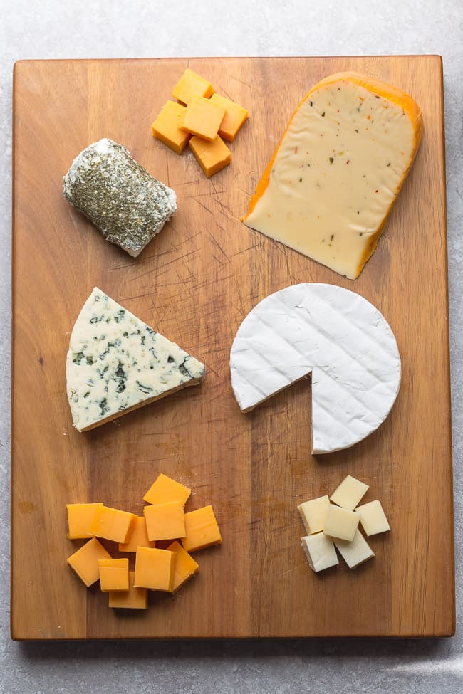 A cutting board with a variety of cheeses on it, including cubes of cheddar, a round of brie, a log of goat cheese, and a slice of blue cheese