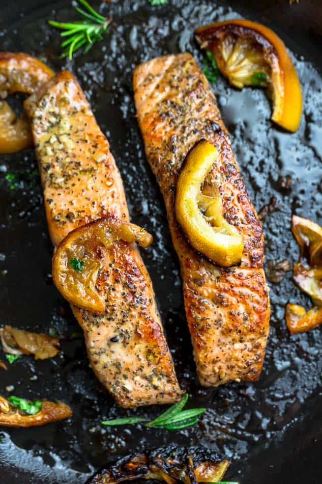 Two pan-fried fish fillets inside of a skillet with oil, herbs and lemon