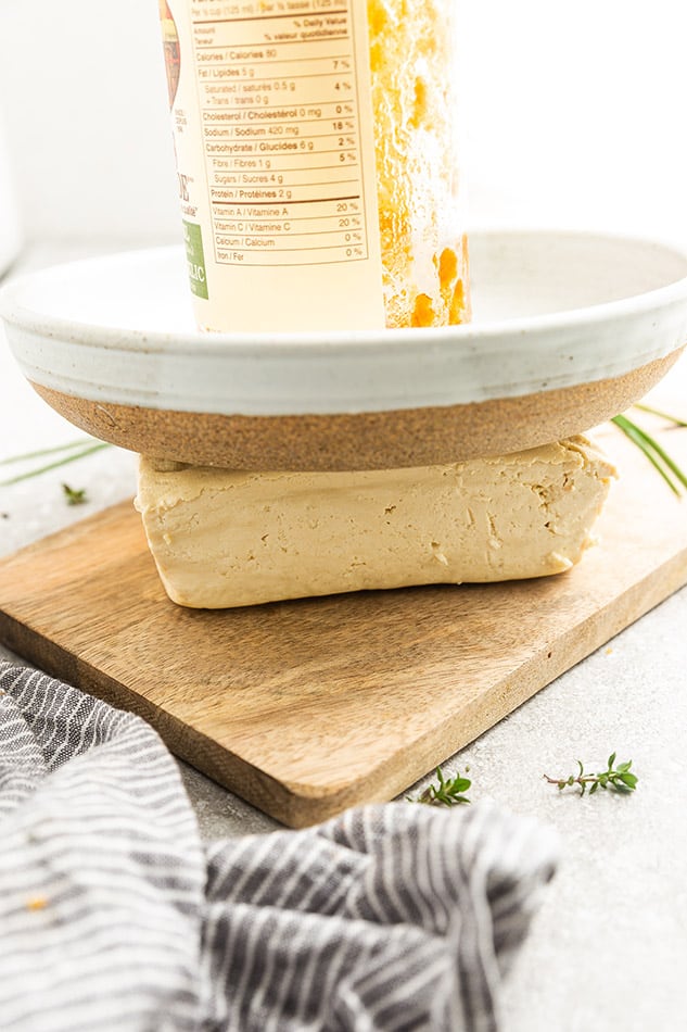 A block of tofu on a wooden cutting board with a heavy plate on top of it as well as a glass can to add extra pressure