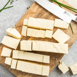 Cubes of uncooked tofu on a wooden cutting board with a sharp chef's knife beside them