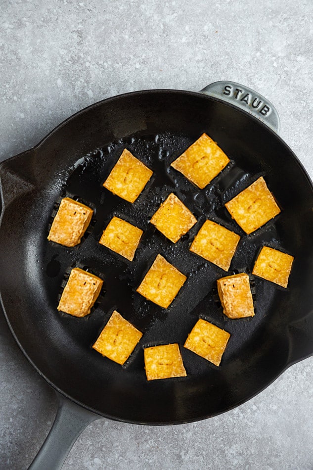 Fourteen pan-fried tofu cubes on a skillet on top of a marble surface