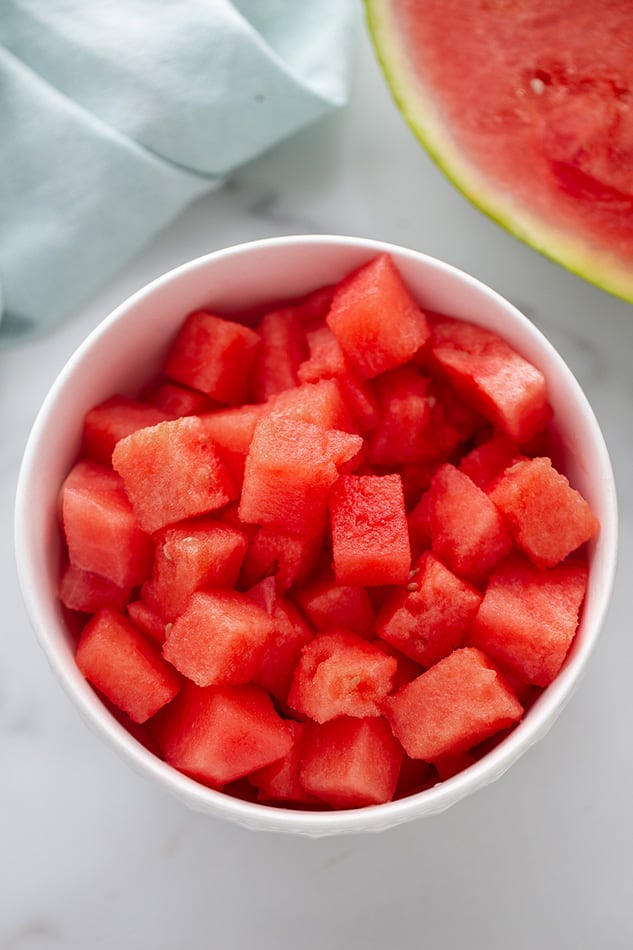 White bowl of watermelon cubes on white surface.