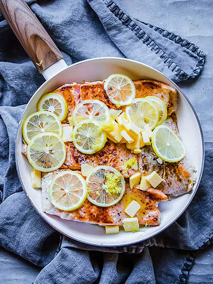 Seared salmon fillets inside of a skillet with butter, lemon juice, parsley, lemon zest and lemon slices