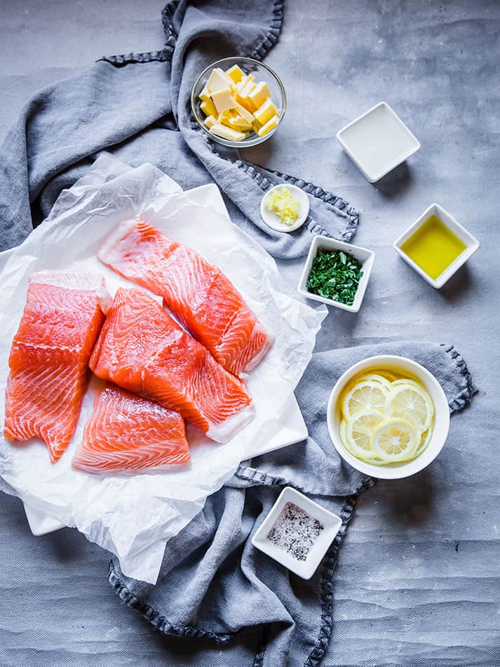 Four descaled fish fillets beside fresh lemon slices, fresh parsley and the rest of the ingredients on a countertop