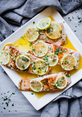A square plate holding four pan-seared salmon fillets topped with fresh lemon slices