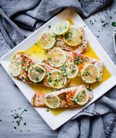 A square plate holding four pan-seared salmon fillets topped with fresh lemon slices