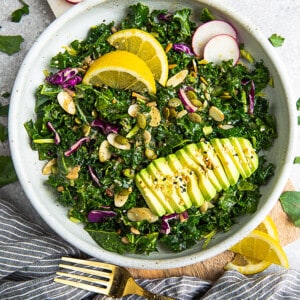 Overhead view of one serving of a kale salad, with kale, avocado and lemon in a white bowl with a gold fork on the side