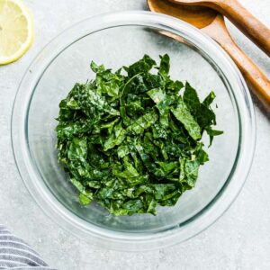 Top view of kale in a clear bowl with caesar dressing on a grey background with wooden spoons