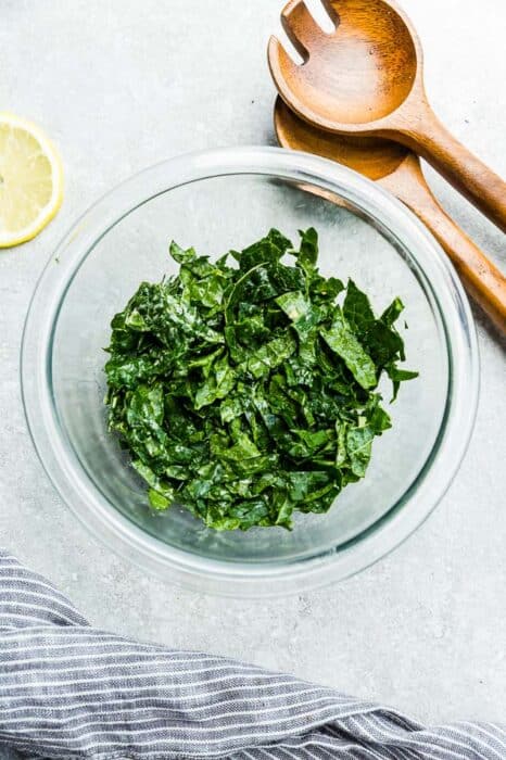 Top view of kale in a clear bowl with caesar dressing on a grey background with wooden spoons