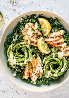Close-up top view of Kale Salad with Chicken and Caesar Dressing in a white bowl on a grey background with lemon slices and a bowl of Caesar dressing