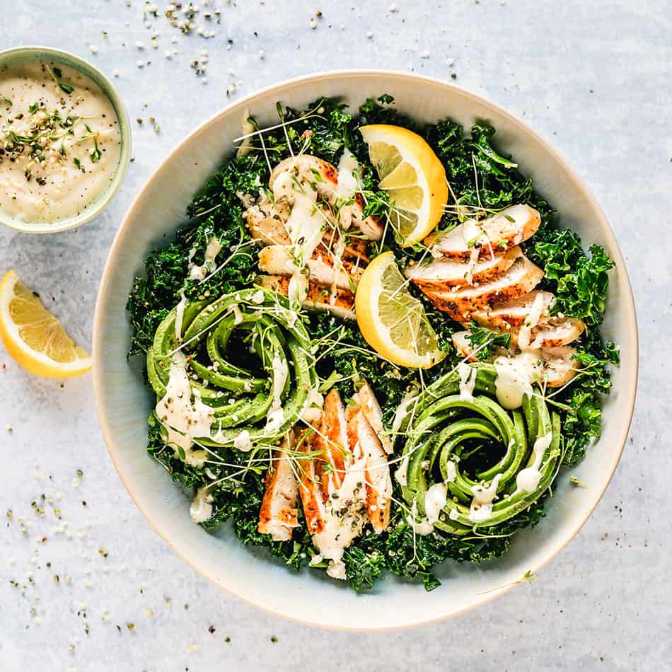 Close-up top view of Kale Salad with Chicken and Caesar Dressing in a white bowl on a grey background with lemon slices and a bowl of Caesar dressing