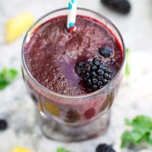 A blackberry kale smoothie in a glass with a blue and white straw and a blackberry on top