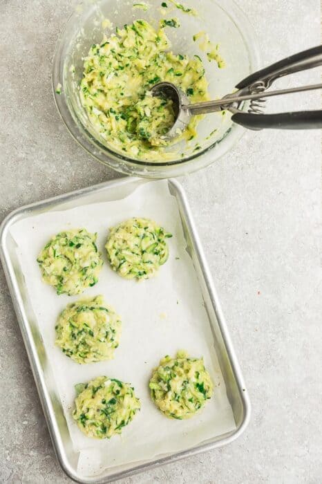 Zucchini fritter batter in a bowl beside a lined baking sheet holding five unbaked fritters