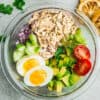 Top view of ingredients to make an avocado chicken salad in a large clear bowl on a grey background
