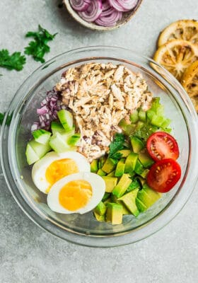 Top view of ingredients to make an avocado chicken salad in a large clear bowl on a grey background