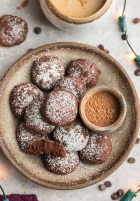 Top view of keto chocolate cookies in a beige bowl with powdered monk fruit sweetener, cocoa powder and christmas lights around the bowl on a grey background