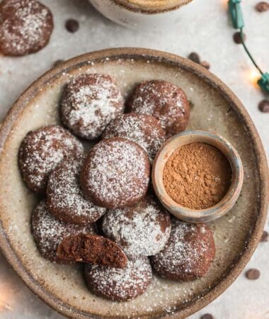 Top view of keto chocolate cookies in a beige bowl with powdered monk fruit sweetener, cocoa powder and christmas lights around the bowl on a grey background